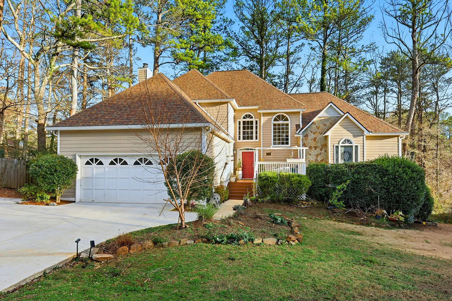 Traditional home with tan siding, a red front door, a two-car garage, and a wooded lot